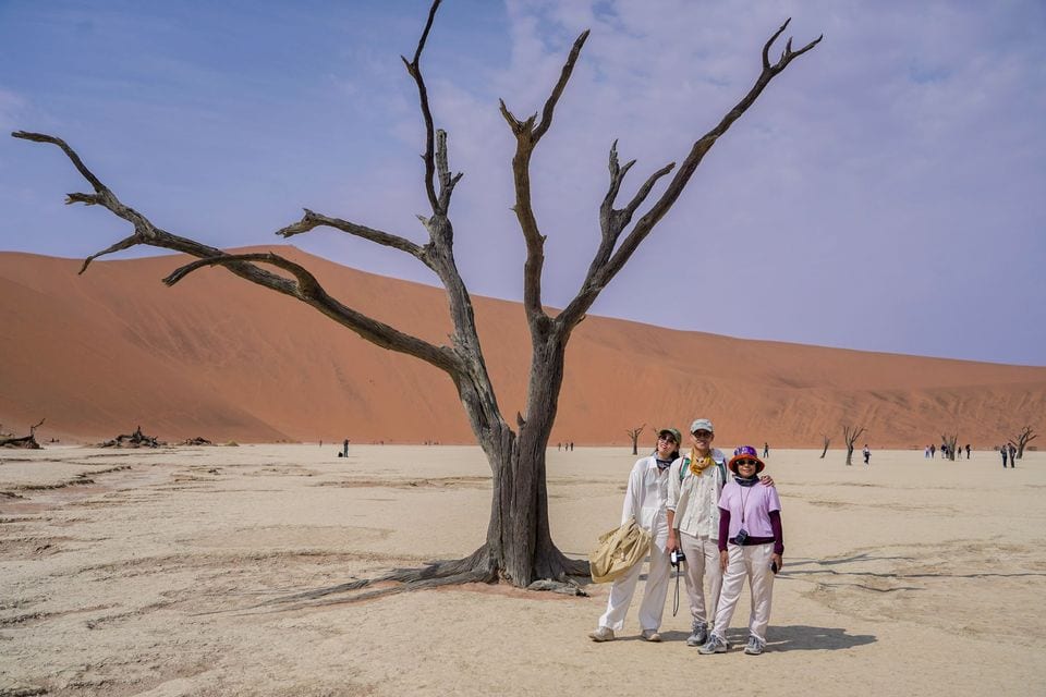 ผู้ร่วมเดินทางสามคนยืนข้างต้นไม้แห้ง โดยมีเนินทรายสีส้มเป็นฉากหลัง